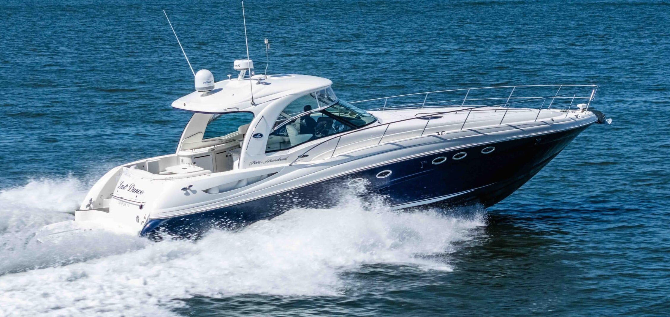White speedboat skimming the blue water with wake spraying behind; a marina and distant hills visible on the horizon.