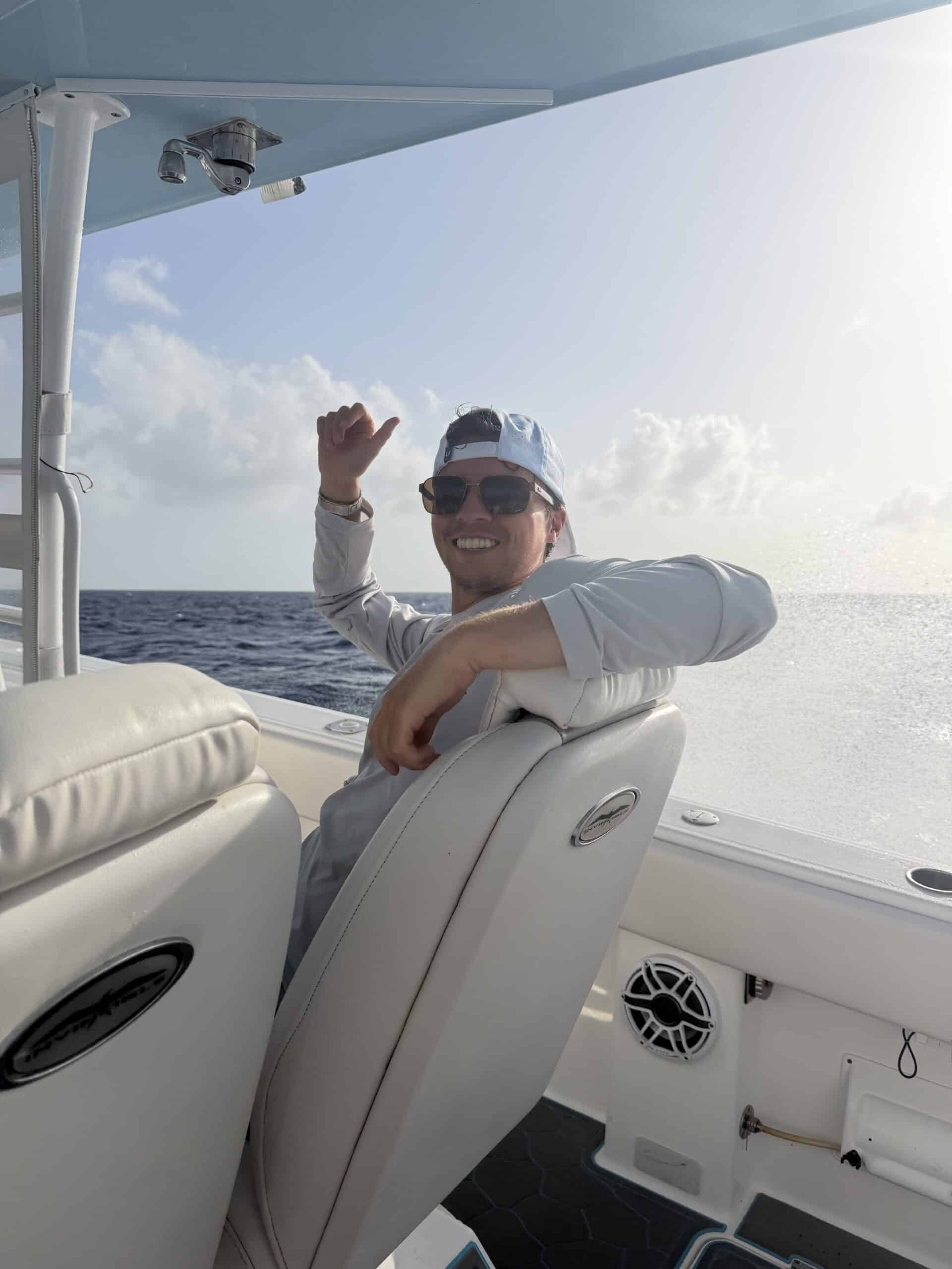 Man wearing a cap and sunglasses sits on a boat seat, smiling at the camera with the ocean in the background.