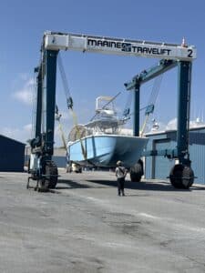 Blue boat suspended in a Marine Travelift hoist between tall blue support towers; a worker stands nearby on the asphalt yard.