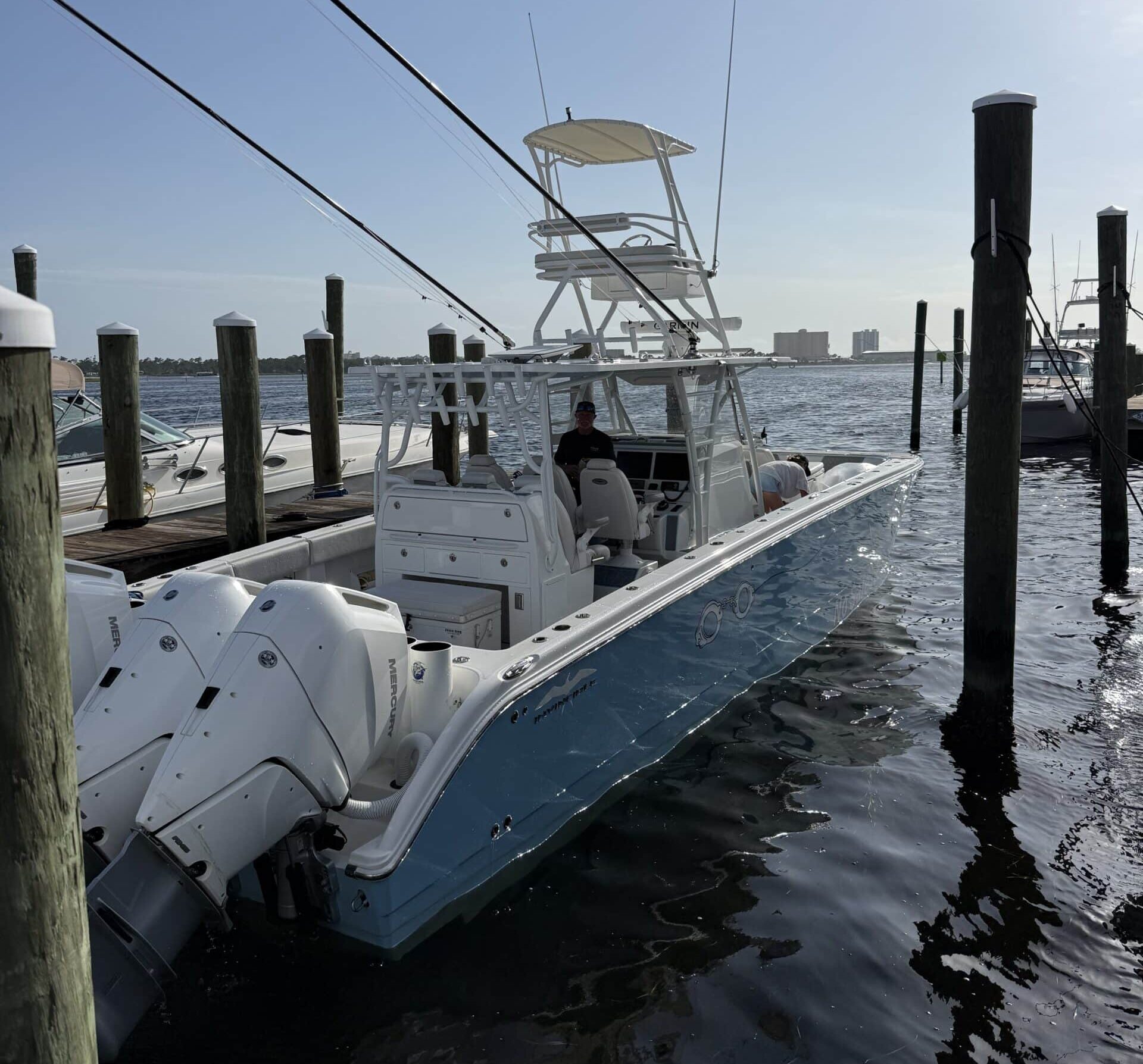 White fishing boat docked at a marina with tall pilings and calm water, blue sky above.
